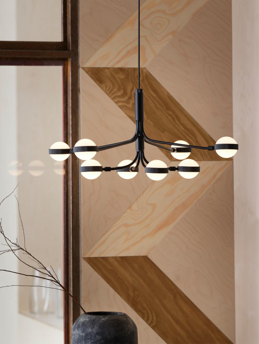 Imaginea arată un candelabru modern cu opt becuri sferice, suspendat de tavan. Fundalul este decorat cu un perete din lemn cu modele geometrice în zigzag. În colțul din stânga jos se află o vază neagră cu crenguțe uscate.