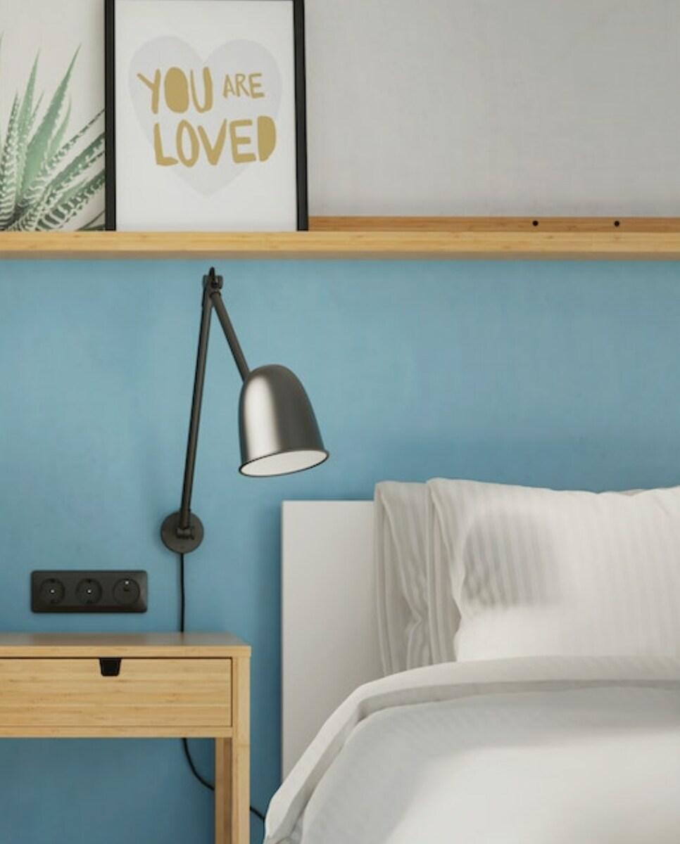 Imaginea arată un colț de dormitor cu un perete albastru, o lampă neagră montată pe perete, o noptieră din lemn cu trei prize electrice și un pat cu lenjerie albă. Pe raftul de deasupra patului este un tablou cu textul "YOU ARE LOVED".