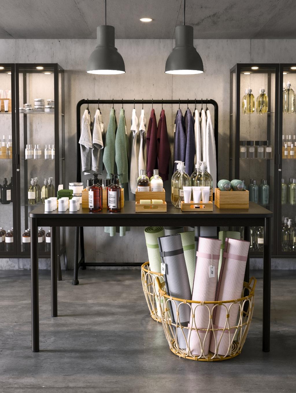 Imaginea arată un magazin de produse pentru îngrijire personală și wellness. Pe masă sunt expuse diverse sticle și recipiente cu produse cosmetice, iar dedesubt sunt coșuri cu covorașe de yoga. În fundal, rafturi cu prosoape și alte produse.