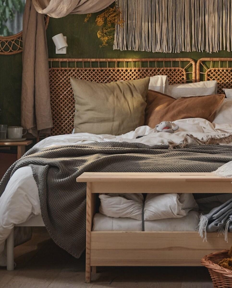 Imaginea arată un pat confortabil, acoperit cu o pătură gri și perne decorative în nuanțe de bej și alb. În fundal, se observă un perete verde decorat cu plante și o perdea albă. Lângă pat este o noptieră cu o cană și alte obiecte mici.