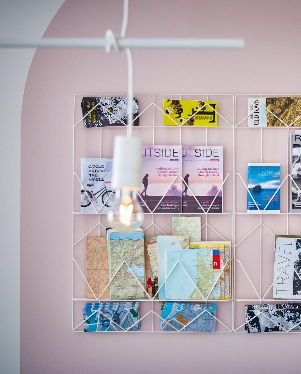 Imaginea arată un perete roz pal cu un suport metalic alb montat pe el. Suportul conține reviste, hărți și broșuri. În prim-plan, este vizibil un bec suspendat de la tavan.