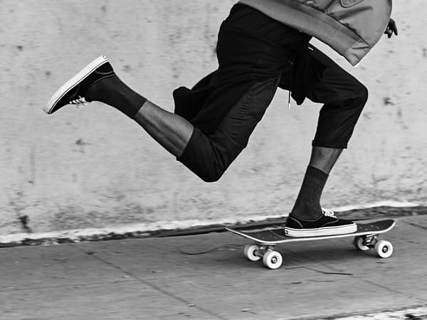 Imaxe recortada en branco e negro onde se ven as pernas dun rapaz en patinete pola beirarrúa.