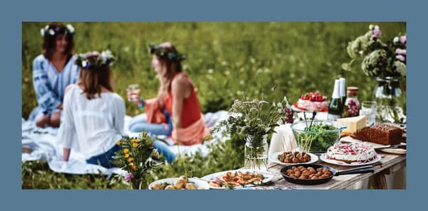 Immagine di tre donne con corone di fiori in testa menre stanno bevendo e parlando sedute su una coperta in un agiardino. Accanto, è allesitito un buffet con cibi e bevande.