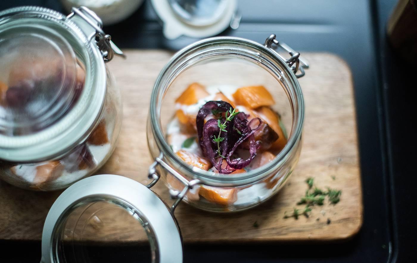 Immagine vista dall'alto di alcuni contenitori KORKEN contenenti alimenti su un tagliere in legno. Uno ha il coperchio aperto e accanto ha delle erbe aromatiche.