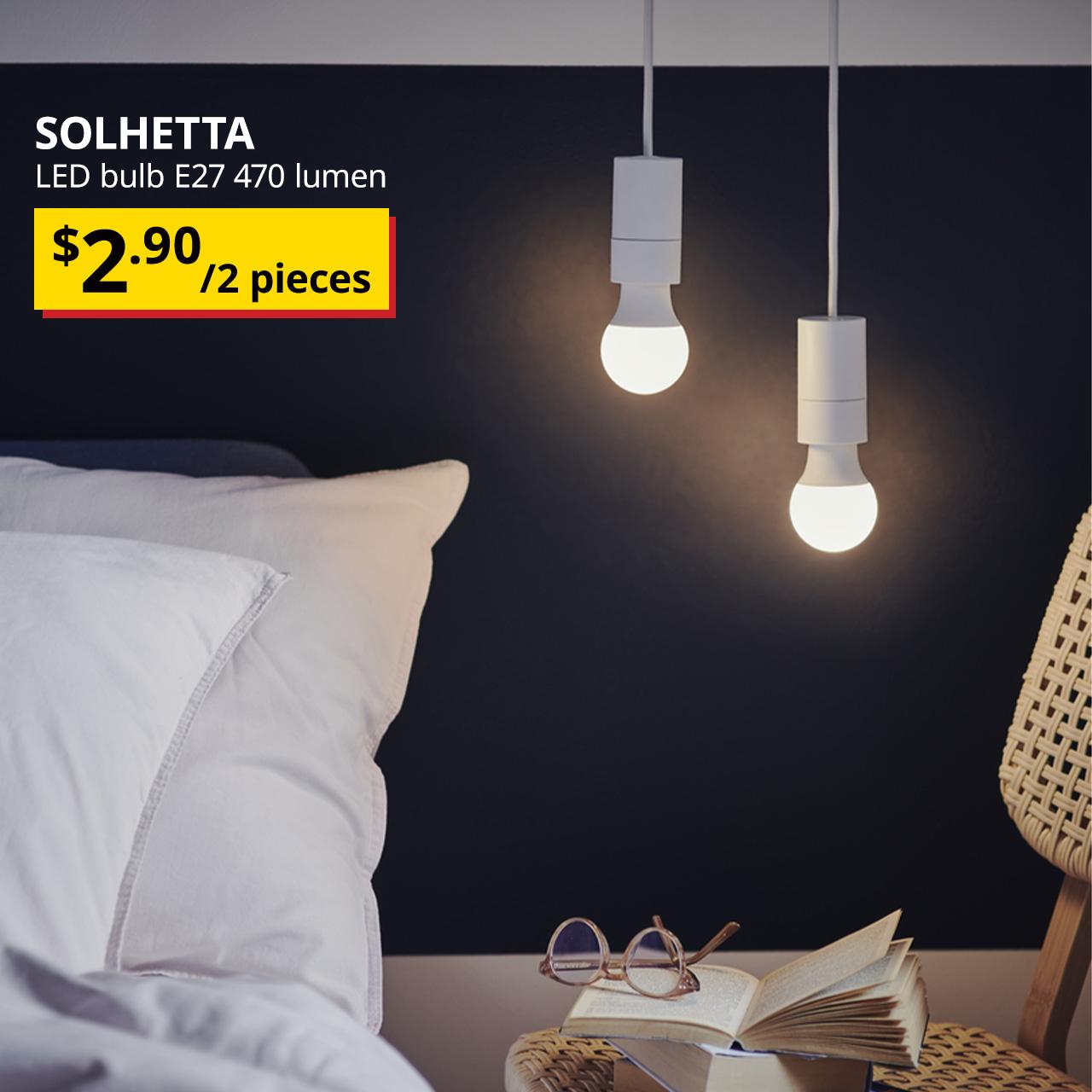 In a bedroom setting, two SOLHETTA bulbs are hung and lit next to the bed. The top left corner of the image, featuring black text that reads: "SOLHETTA, LED bulb E27 470 lumen" with the BTI price in yellow/red and black text: $2.90/2pieces.