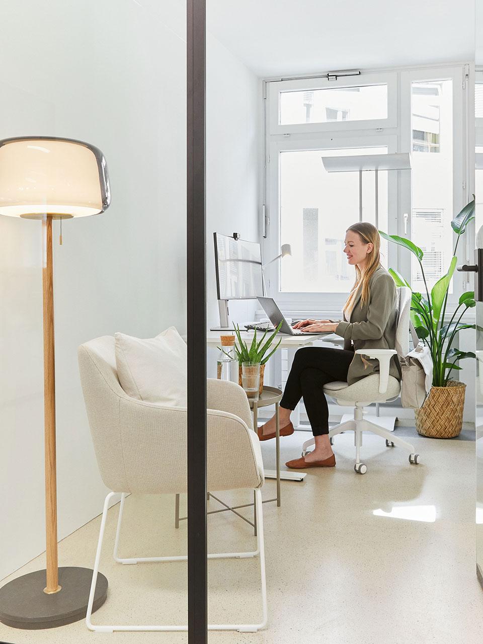 In a bright office space furnished with armchairs and plants, a person sits on an office chair working at a desk.