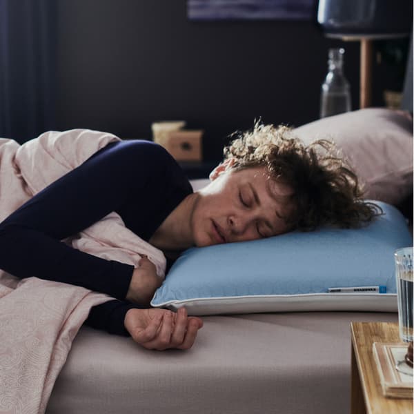 In a dark bedroom, a person in blue pyjamas in bed with a KVARNVEN ergonomic pillow is sleeping on their side.