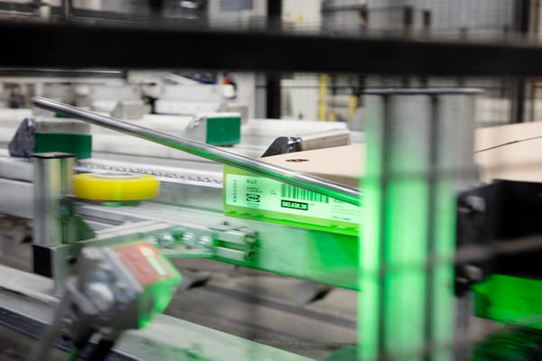 In a factory environment, flat packages travel along a conveyor belt. A green light beam scans barcodes on the packages.