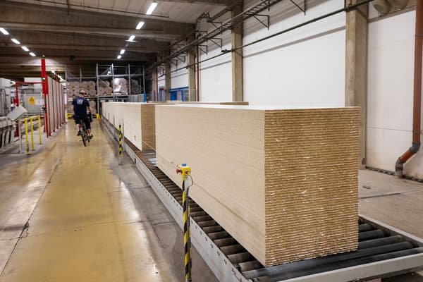 In a factory premises, particleboards are stacked on top of each other. A factory worker cycles by.