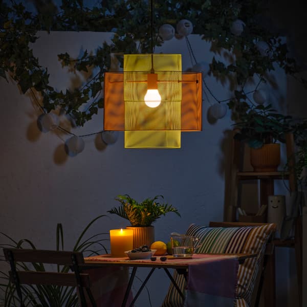 In a green outdoor space, an orange and light green SOLVINDEN pendant lamp casts a warm glow over a table and two chairs.