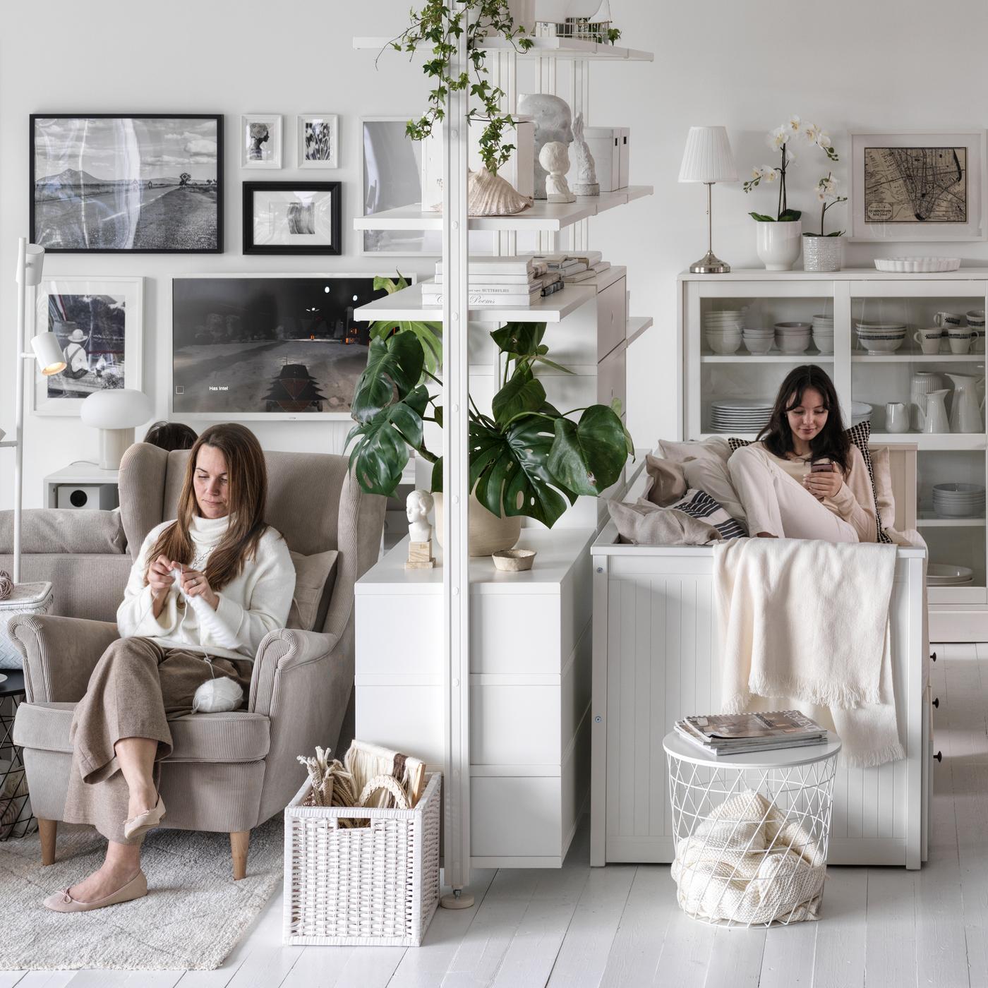In a living room with white walls and floor, two people sit either side of a white ELVARLI open storage combination.