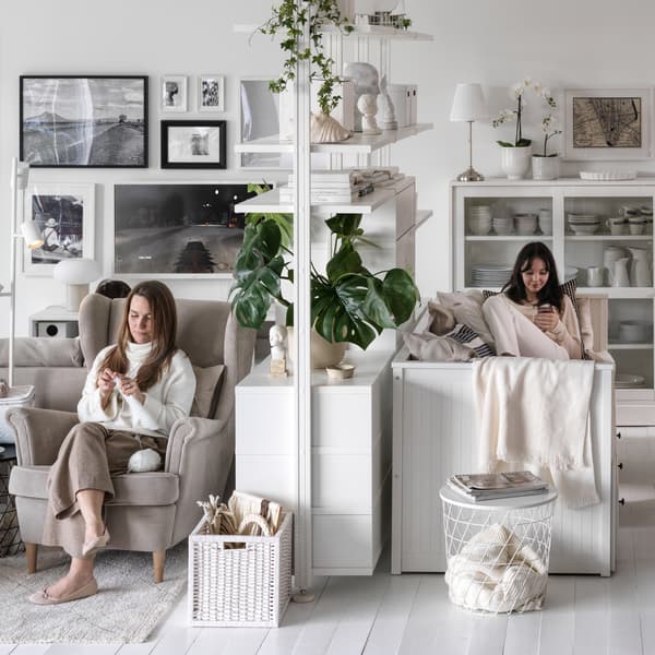In a living room with white walls and floor, two people sit on either side of a white ELVARLI open storage combination.