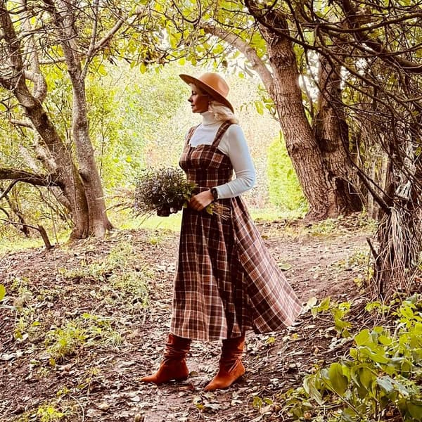 In a side view, a woman wearing a brown, checkered dress, brown boots, and a wide-brimmed hat stands in an alley, holding a bouquet of flowers.				