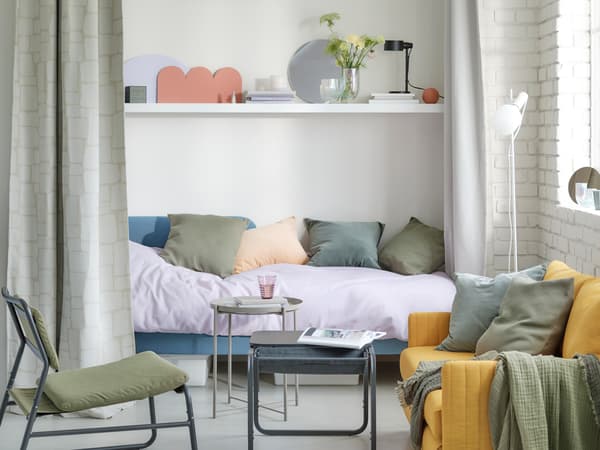 In a small living room is a medium blue BLÅKULLEN bed with corner headboard filled with pillows in different pastel colours.
