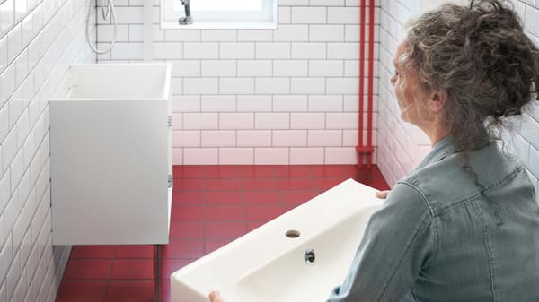 In a white and red tiled bathroom, a woman holds a white wash basin and looks at a white base cabinet fixed to the wall.