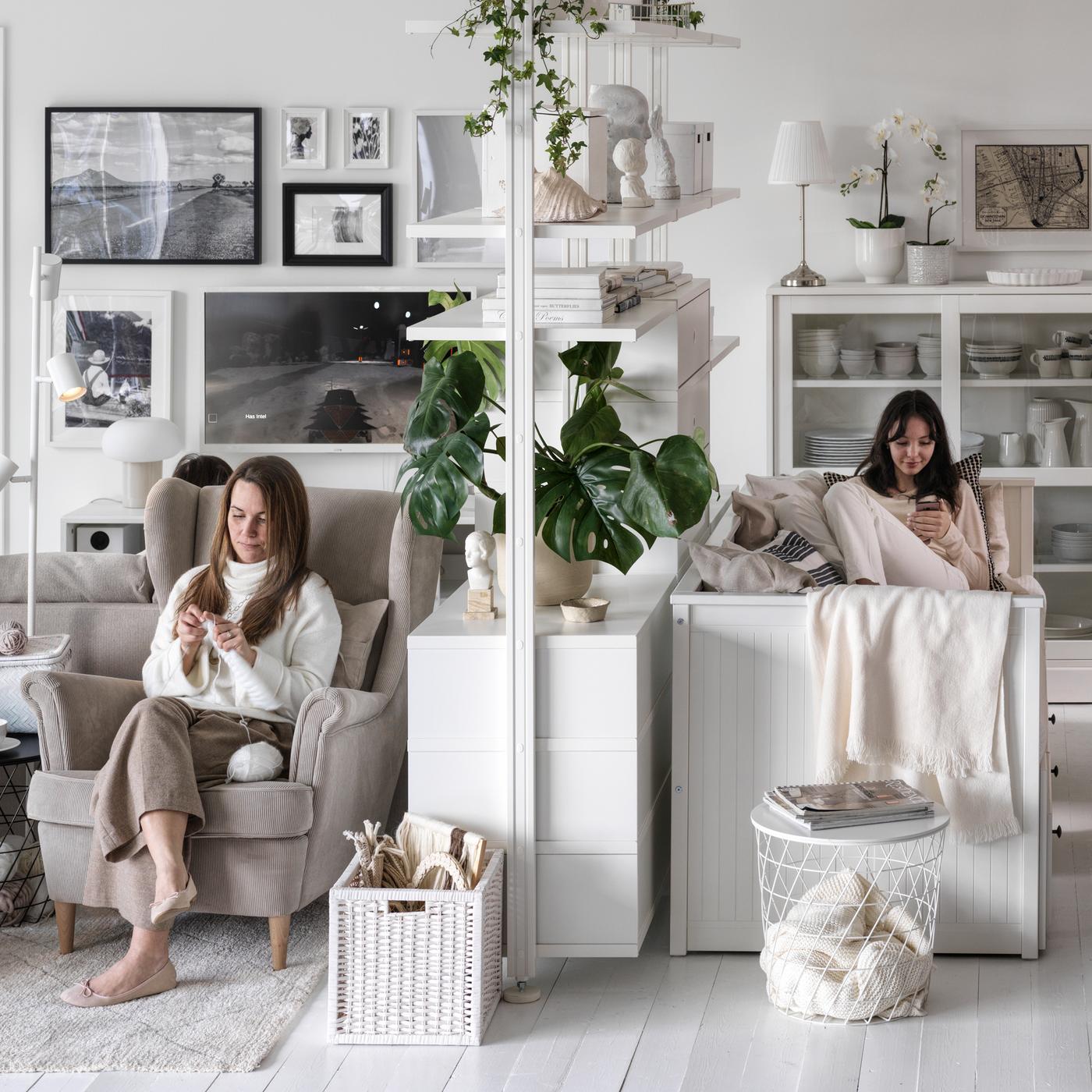 In een woonkamer met witte wanden en vloer zitten twee mensen aan beide kanten van een witte ELVARLI open opbergcombinatie.