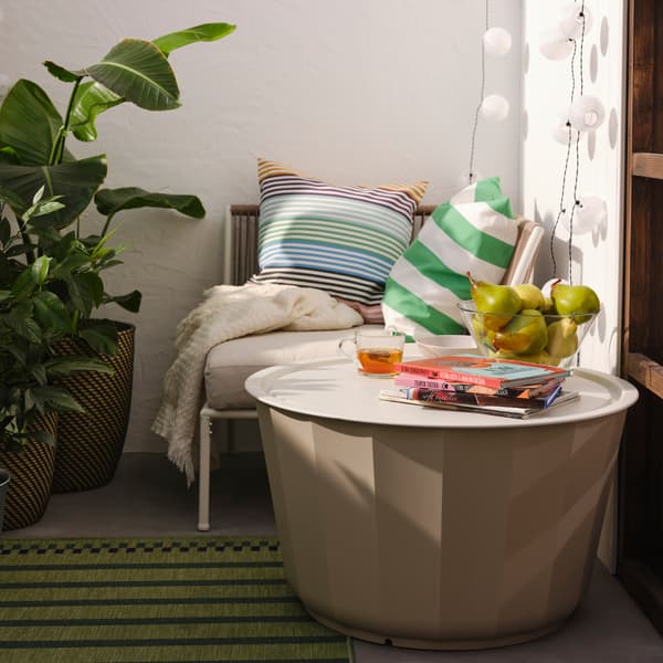 In einem Aussenbereich ist ein beigefarbener LÅGASKÄR Couchtisch für draussen auf einem gestreiften Teppich in Grün/Lila neben einem Sitzeckelement in Weiss/Beige mit Kissen und einem Plaid zu sehen.
