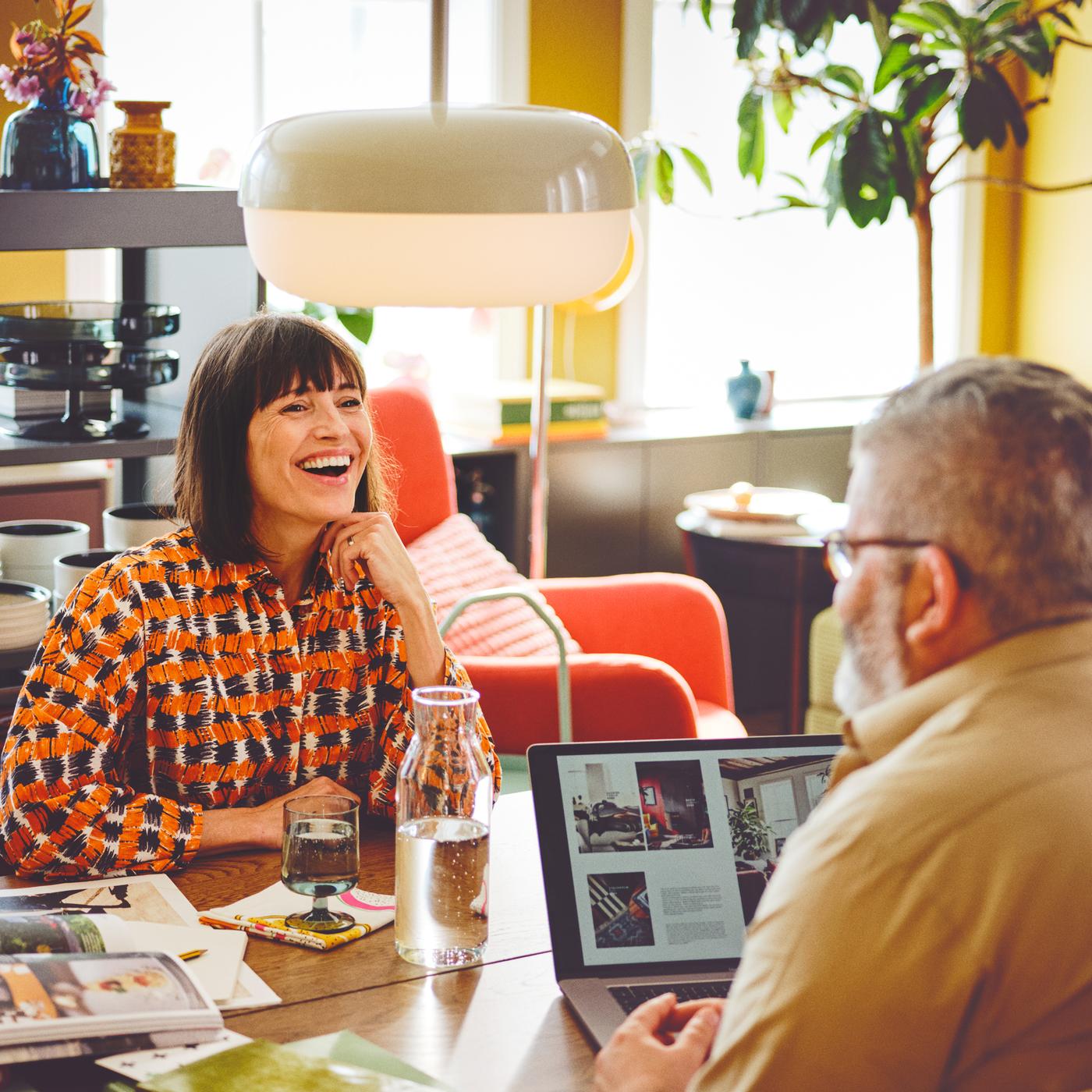 In einem farbenfrohen Raum sitzen sich zwei Personen an einem Tisch gegenüber. Auf dem Tisch befinden sich ein Laptop und eine Karaffe. Darüber hängt eine BLÅSVERK Hängeleuchte.