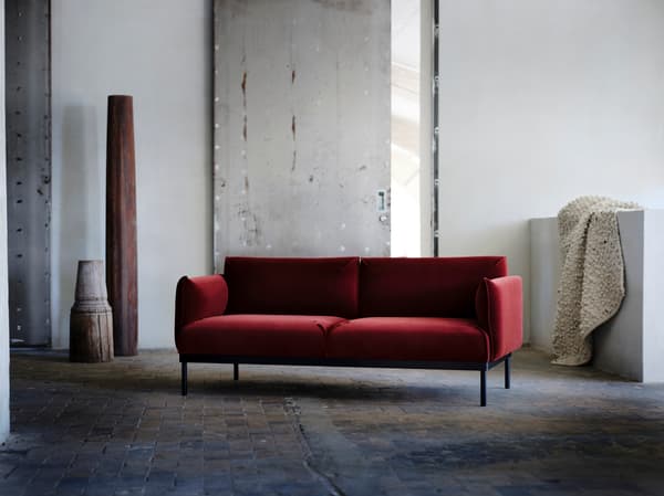 In einem hellen Raum mit grauen Wänden und Steinboden steht das rote APPLARYD 2er-Sofa von IKEA mit einem flach gewebten SVÄRDBORG Teppich.