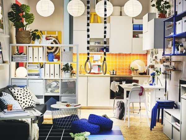 In einem hellen Wohnbereich sind eine weiße Küchenzeile, ein KALLAX Regal, ein Klapptisch mit Stühlen, eine Couch und viele weitere Möbelstücke und Wohnaccessoires zu sehen.