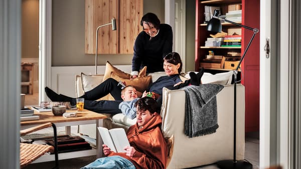 In einem Wohnzimmer befinden sich vier Personen auf und um ein BACKSÄLEN 3er-Sofa, das vor einem rot und hellbraun lasiertem HEMNES Bücherregal steht.