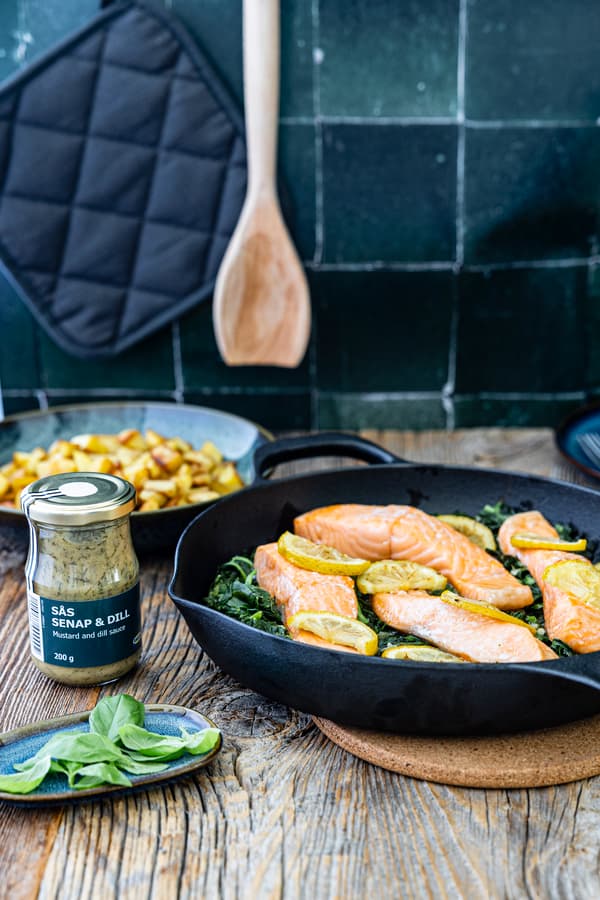 In einer Pfanne befinden sich frischer Spinat, Lachfilets und Zitronenscheiben. Drumherum sind Küchenutensilien zu sehen.