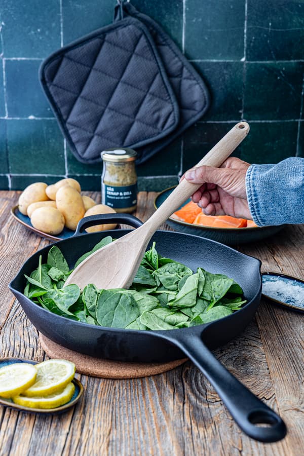 In einer Pfanne befindet sich frischer Spinat, der mit einem Holzlöffel gerührt wird.