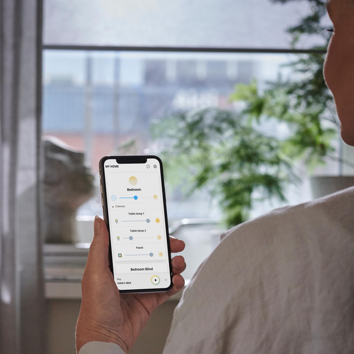 In front of a window, a person’s hand holds a smart phone with an app on the screen to control electric lighting and blinds.