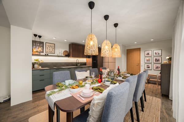 In the foreground, a covered dining table surrounded by 8 gray fabric-covered chairs with woven lampshades hanging above it. In the background, a gray-green kitchen.