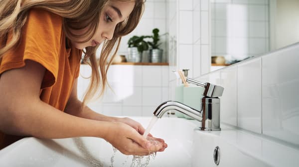 In un bagno piastrellato di bianco, un bambino in piedi con lunghi capelli castani si lava le mani nel lavabo.
