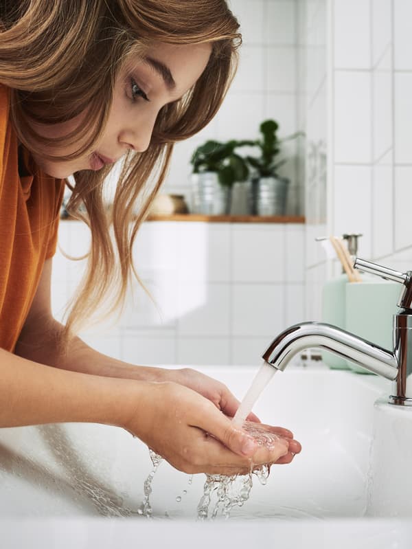 In un bagno piastrellato di bianco, una ragazza in piedi con lunghi capelli si lava le mani nel lavabo.
