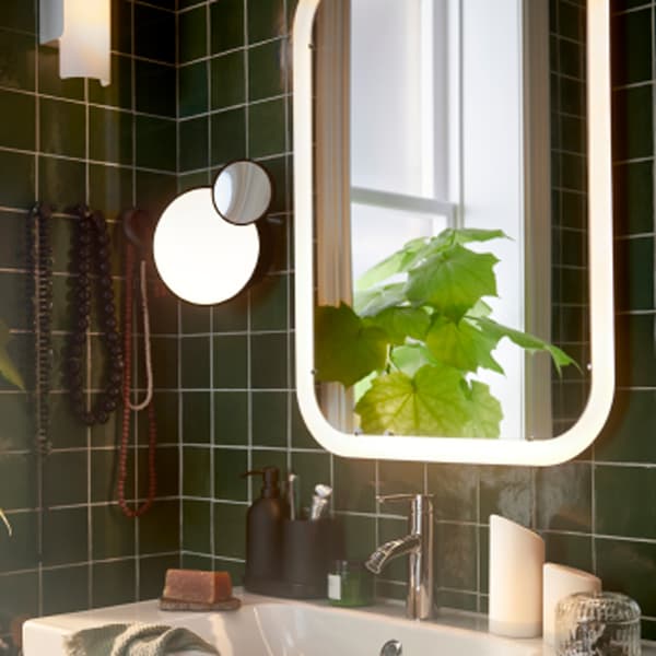 In un bagno, uno specchio bianco con illuminazione integrata posizionato sopra a un mobile per lavabo, due ceri a LED e una pianta in vaso.