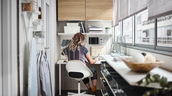 In un balcone, una persona seduta in una sedia girevole beige/bianca vicino a uno scaffale con piano tavolo BOAXEL/LAGKAPTEN bianco - IKEA.