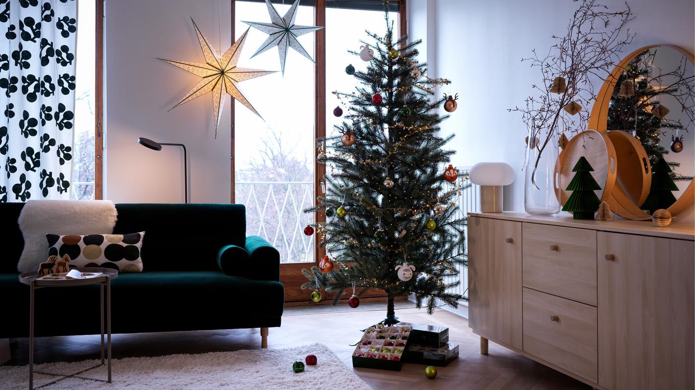 In un soggiorno, albero di Natale VINTER 2021 decorato con palline accanto a un divano verde e decorazioni a sospensione a forma di stella.