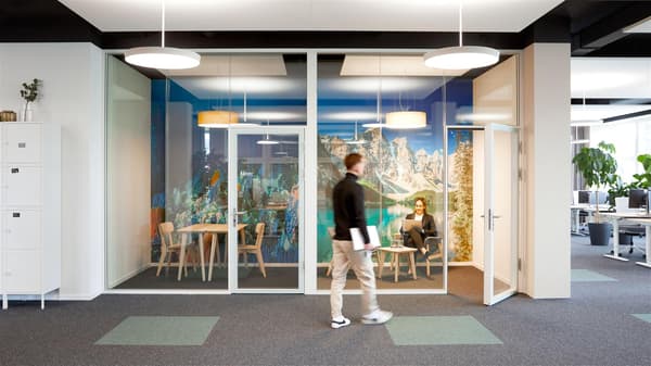 In un ufficio open space, una persona passa davanti a una piccola sala riunioni vetrata, con carta da parati panoramica sulla parete di fondo