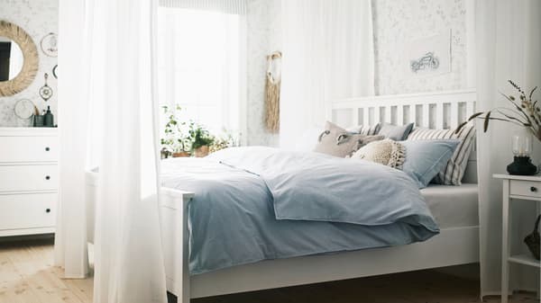 In una camera luminosa, letto HEMNES bianco circondato da tende sottili SILVERLÖNN bianche, con cuscini e biancheria blu - IKEA.