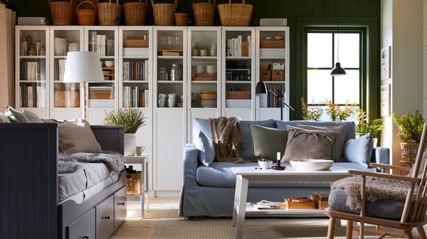 In witte BILLY boekenkasten met paneel-/vitrinedeuren zitten boeken, bekers en schalen. Een blauwe zitbank en witte salontafel staan voor de kasten.