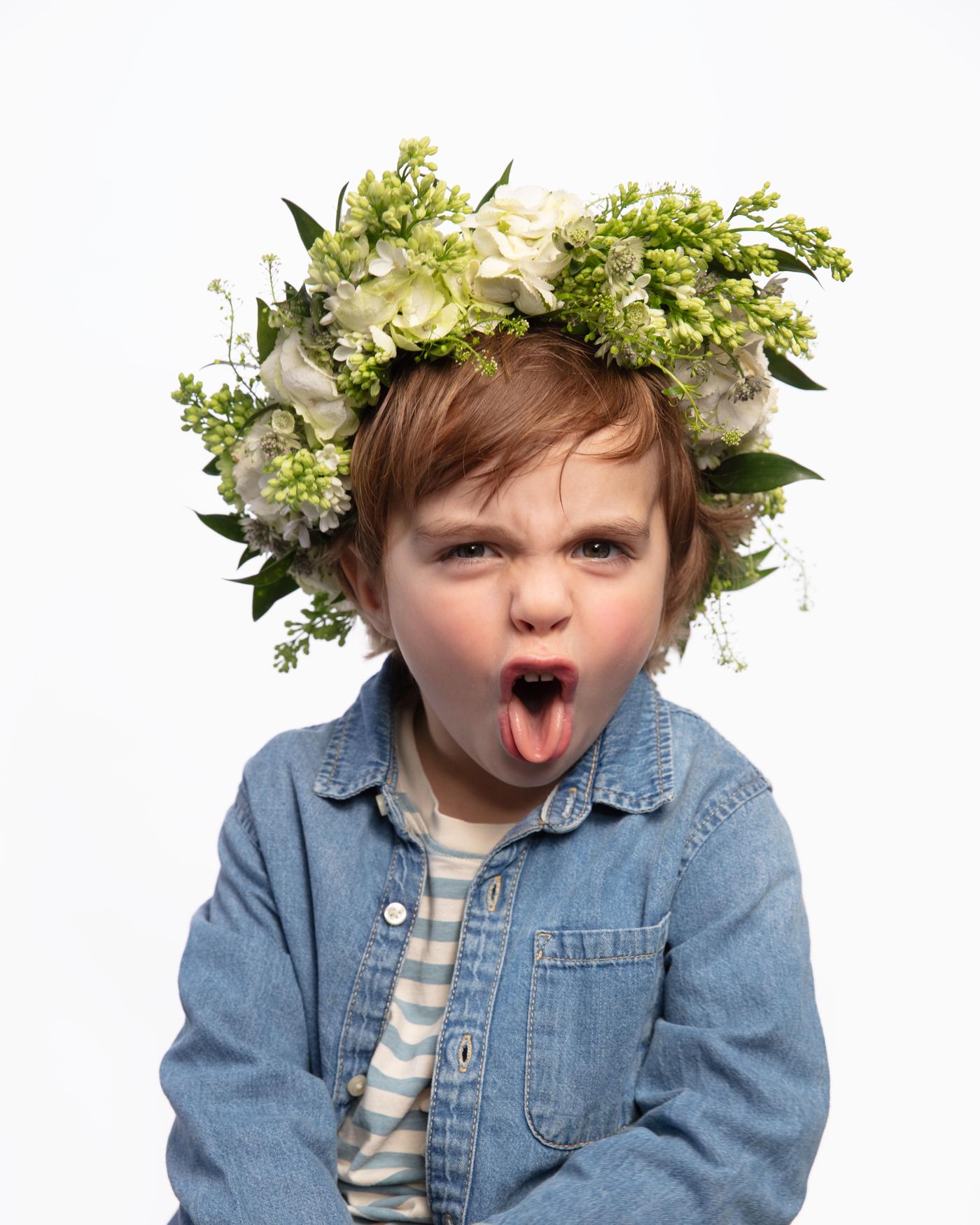 Infant fent ganyota amb la llengua fora, camisa texana i corona de flors blanques i verdes al cap.