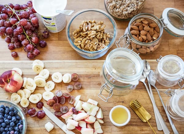 Ingredientes para o pequeno-almoço, incluindo fruta cortada, nozes e recipientes de vidro, espalhados numa bancada em madeira.