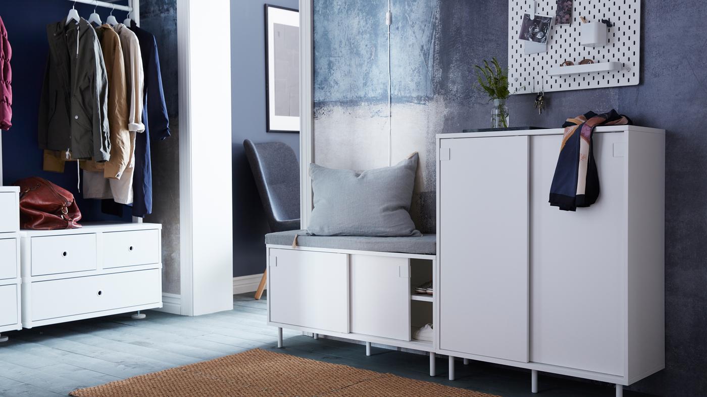 Ingresso con panca con vano contenitore MACKAPÄR e cuscino grigio, vicino a un mobile MACKAPÄR su cui sono appoggiati un vaso e una sciarpa. Sopra il mobile un pannello portaoggetti SKADIS. Illuminata dalla luce del sole, un sistema componibile ELVARLI con due cassettiere, bastone appendiabiti e ripiani 