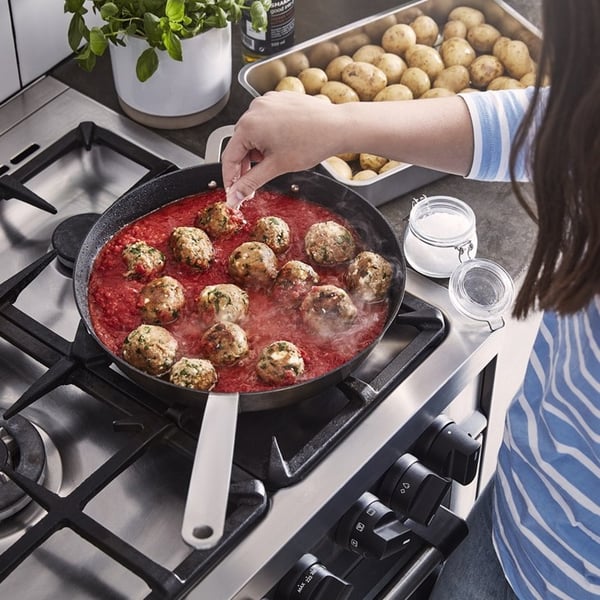 Inimene raputab pliidil punases kastmes küpsevatele lihapallidele maitseaineid; läheduses on ahjuplaat kartulitega ja muud toidutegemiseks vajaminevad koostisosad.