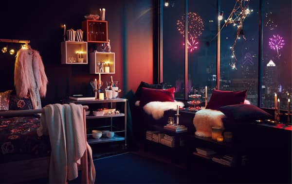 Intérieur d’appartement avec décorations et éclairage à la fête ; feux d’artifice à l’horizon d’une ville visibles à travers de hautes fenêtres.