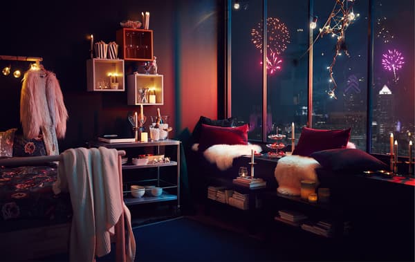 Intérieur d’un appartement avec des décorations de fête élégantes et beaucoup de lumières; on peut voir à travers les larges fenêtres un feu d’artifice au-dessus d’un paysage urbain.