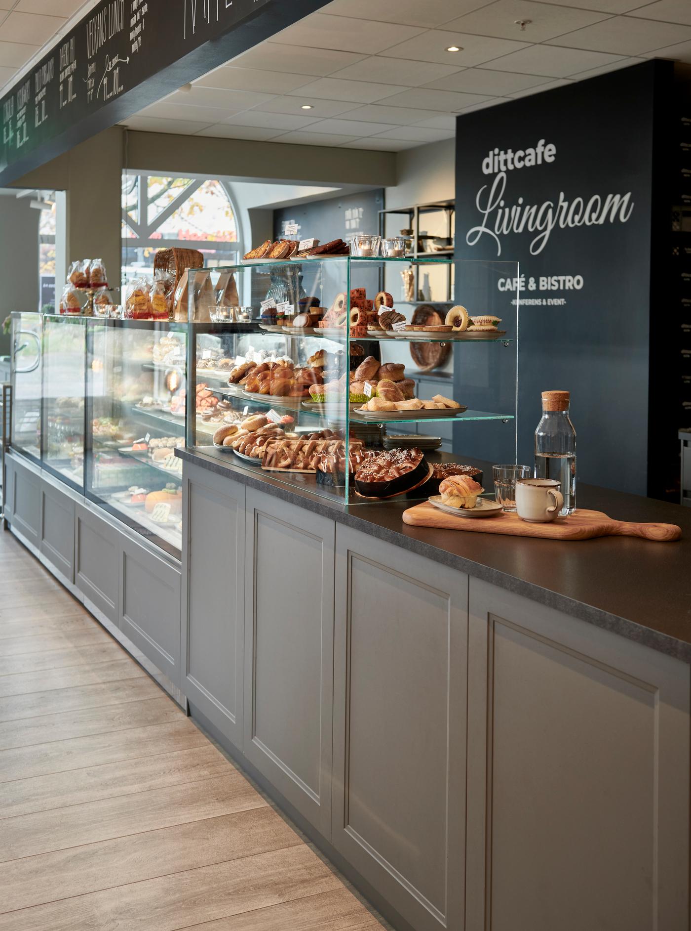 Interiör från caféet 'dittcafe Livingroom' med en glasmonter fylld med bakverk, bröd och desserter. Modern inredning med trädetaljer och meny på väggen ovanför.
