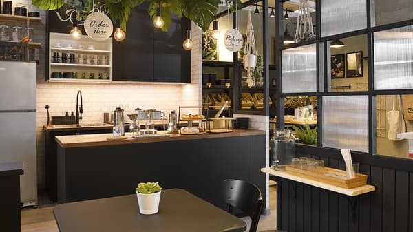 Interior of a café – a black kitchen island with coffee jugs and bread as the order and collection point,  and the dining area with some tables and chairs.