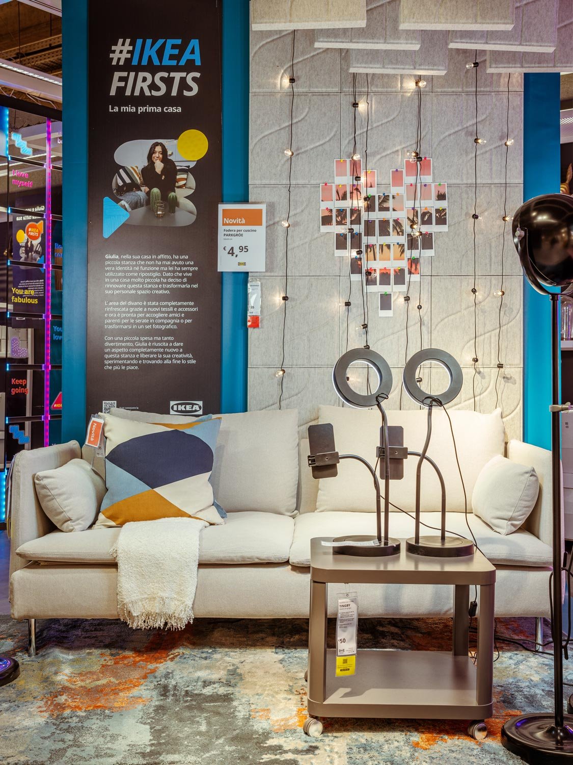 Interno di un negozio IKEA con un divano decorato con cuscini. Ci sono anche supporti per smartphone, lampade posti su di un tavolino. C'è anche un cartellone con la scritta #IKEAfirsts.