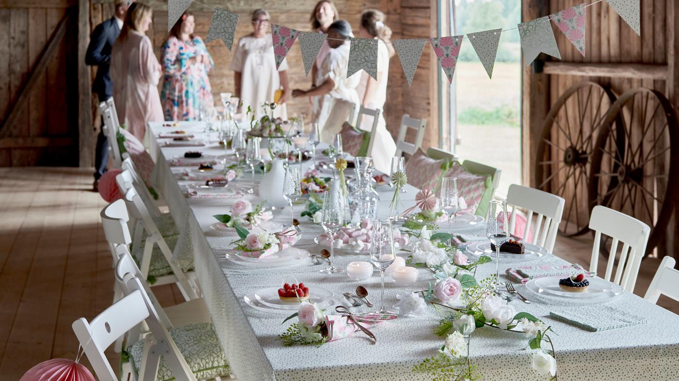 Într-un hambar, este aranjată o masă pentru o petrecere cu pahare, veselă și decorațiuni din colecția festivă INBJUDEN.