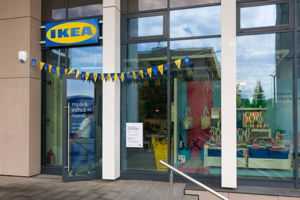 Intrarea unui magazin IKEA cu semn albastru și galben; în față sunt stegulețe decorative, iar în interior se văd perne colorate și coșuri suspendate.