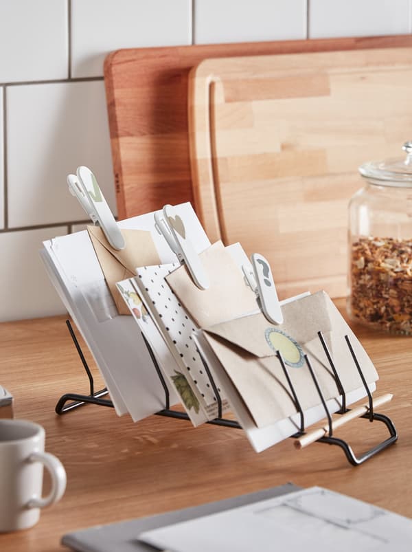 Între accesorii de bucătărie împrăștiate, un suport de farfurii RINNIG așezat orizontal este folosit ca suport pentru plicuri și hârtii.