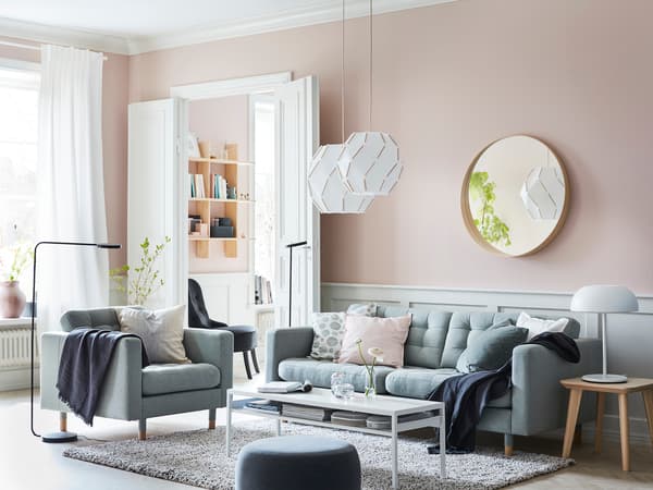 淡いピンクの壁にグレーソファを置いた北欧風リビング。白いカーテンと丸いミラーで柔らかな雰囲気。